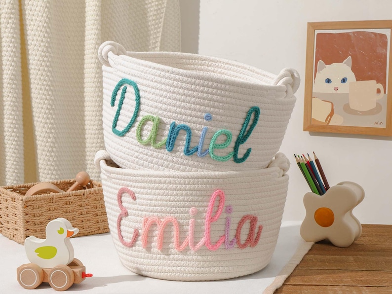May include: Two white rope storage baskets with colourful embroidered names, "Daniel" and "Emilia". The baskets are stacked on top of each other.