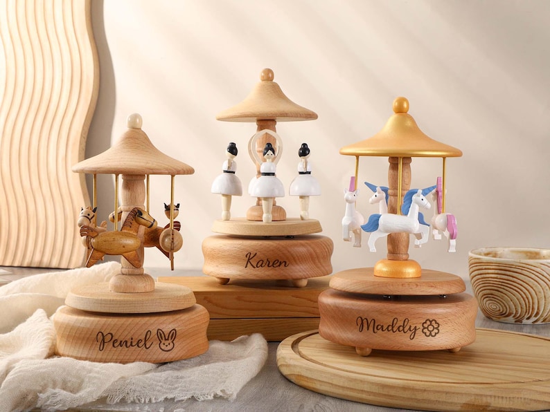 May include: Three wooden music boxes with carousel designs. The first music box features a carousel with horses and the name "Peniel" engraved on the base. The second music box features a carousel with ballerinas and the name "Karen" engraved on the base. The third music box features a carousel with unicorns and the name "Maddy" engraved on the base.