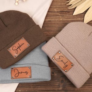 May include: Three knitted beanies in brown, beige, and blue with leather patches that have names written on them. The names are "Sophia", "Emily", and "James".