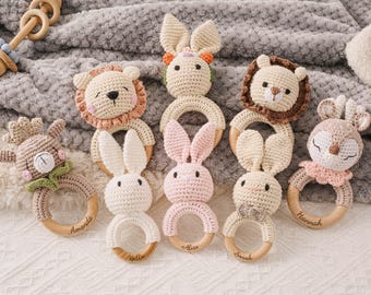 May include: A collection of handmade crochet baby rattles in various animal shapes, including a lion, bunny, and deer. Each rattle features a wooden ring and is personalized with a name. The rattles are in soft, neutral colors.