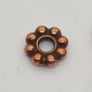 May include: A copper-colored metal bead with a round hole in the center. The bead is shaped like a flower with eight rounded petals.