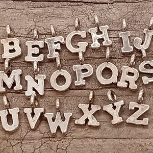May include: Silver alphabet letter charms, A through Z, with a slightly raised, vintage-style font. The charms are arranged in alphabetical order on a wooden surface.