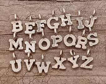 May include: Silver alphabet letter charms, A through Z, with a slightly raised, vintage-style font. The charms are arranged in alphabetical order on a wooden surface.