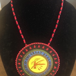 May include: A beaded necklace with a large circular pendant. The pendant features a yellow center with a brown emblem, surrounded by blue, green, and black beaded rings. The necklace has a red and black beaded chain.