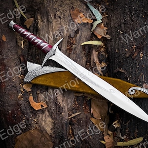 May include: A silver and red dagger with an ornate handle and blade, resting on a wooden surface. The dagger is sheathed in a gold-colored scabbard with silver accents. The blade has decorative etchings.