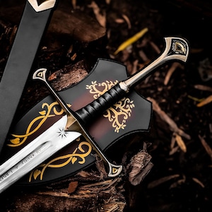 May include: A silver-toned metal sword with a black leather-wrapped grip and a crossguard featuring gold-colored accents. The blade is engraved with runic characters and a star symbol. It is accompanied by a black scabbard and a black display plaque.