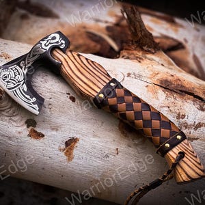 May include: A small, hand-forged axe with a black and silver blade featuring a Celtic knot design. The axe has a wooden handle wrapped in brown leather with a braided pattern and secured with brass rivets.