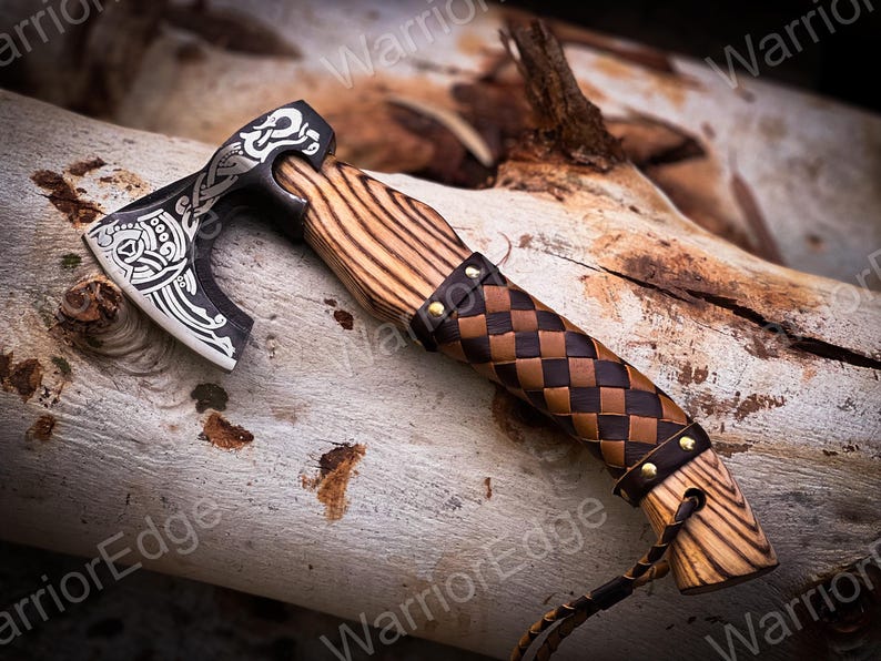 May include: A small, hand-forged axe with a black and silver blade featuring a Celtic knot design. The axe has a wooden handle wrapped in brown leather with a braided pattern and secured with brass rivets.