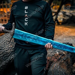 Pode incluir: Uma espada de fantasia com uma l&acirc;mina azul e uma guarda ornamentada. A l&acirc;mina apresenta um desenho de folhas. A guarda &eacute; de cor bronze. A pessoa na imagem est&aacute; vestindo um moletom preto com a palavra "BLACK" em letras brancas.