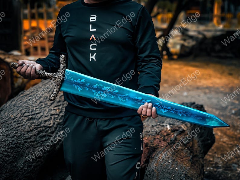 Pode incluir: Uma espada de fantasia com uma l&acirc;mina azul e uma guarda ornamentada. A l&acirc;mina apresenta um desenho de folhas. A guarda &eacute; de cor bronze. A pessoa na imagem est&aacute; vestindo um moletom preto com a palavra "BLACK" em letras brancas.
