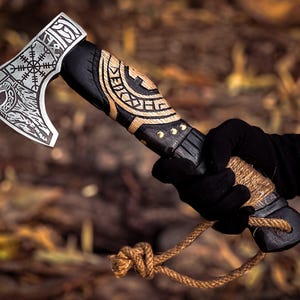 May include: A black and brown axe with a wooden handle and a rope attached to the end. The axe head has a Viking design with a runic symbol and a wolf's head.