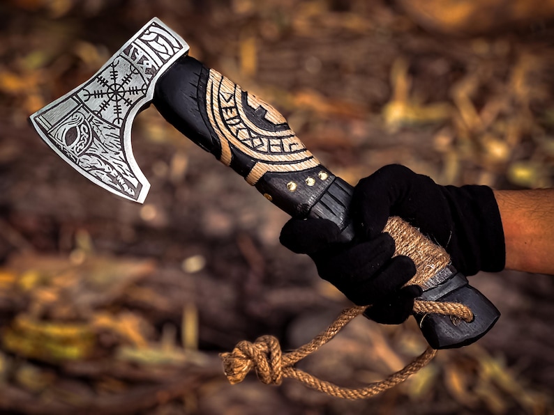 May include: A black and brown axe with a wooden handle and a rope attached to the end. The axe head has a Viking design with a runic symbol and a wolf's head.