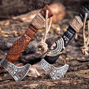 May include: Two miniature Viking axes with intricate carvings on the handles. The axes are made of wood and metal and have a brown and black finish. The axes are tied together with twine and have a decorative knot at the end.