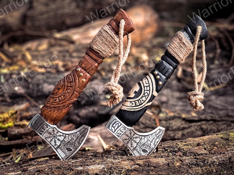 May include: Two miniature Viking axes with intricate carvings on the handles. The axes are made of wood and metal and have a brown and black finish. The axes are tied together with twine and have a decorative knot at the end.