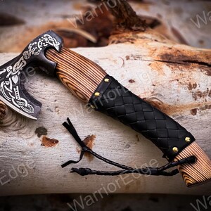 May include: A black and silver axe with a wooden handle wrapped in black leather. The axe has a decorative design on the blade and a leather strap attached to the handle.