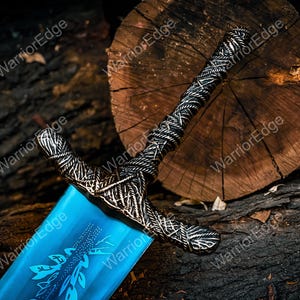 Pode incluir: Espada decorativa com uma l&acirc;mina azul e uma guarda prateada ornamentada com desenhos intrincados. A guarda apresenta uma pega texturizada. A l&acirc;mina apresenta um padr&atilde;o de folhas. A espada repousa sobre uma superf&iacute;cie de madeira escura.
