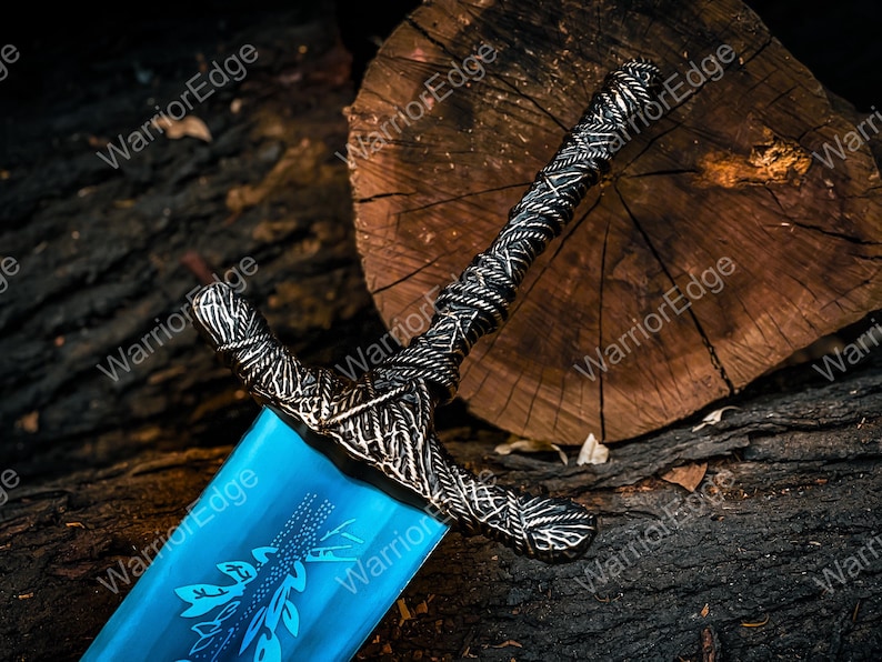 Pode incluir: Espada decorativa com uma l&acirc;mina azul e uma guarda prateada ornamentada com desenhos intrincados. A guarda apresenta uma pega texturizada. A l&acirc;mina apresenta um padr&atilde;o de folhas. A espada repousa sobre uma superf&iacute;cie de madeira escura.
