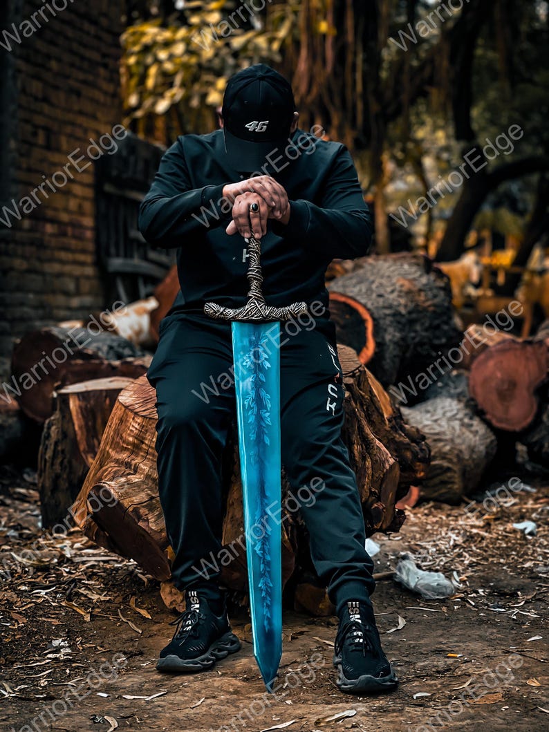 Pode incluir: Uma espada azul e prateada com uma empunhadura ornamentada. A l&acirc;mina tem um desenho de folhas. A pessoa est&aacute; usando um bon&eacute; preto com o n&uacute;mero 46, uma camisa preta de manga comprida e cal&ccedil;as pretas. A espada est&aacute; apoiada em um tronco.