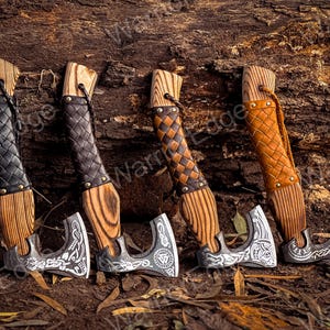May include: Four small hand-crafted axes with wooden handles and leather wrappings. The axes have intricate metal designs and are displayed against a rustic wooden background.