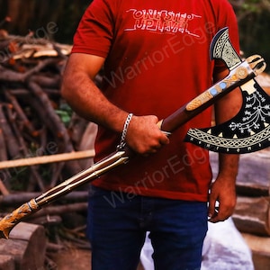 May include: A black axe with gold and silver accents. The axe head has a decorative design with runes and symbols. The axe handle is made of wood and has a leather wrap. The axe is held by a person wearing a red shirt with the text "One Step Up Level" and "Warrior Edge".