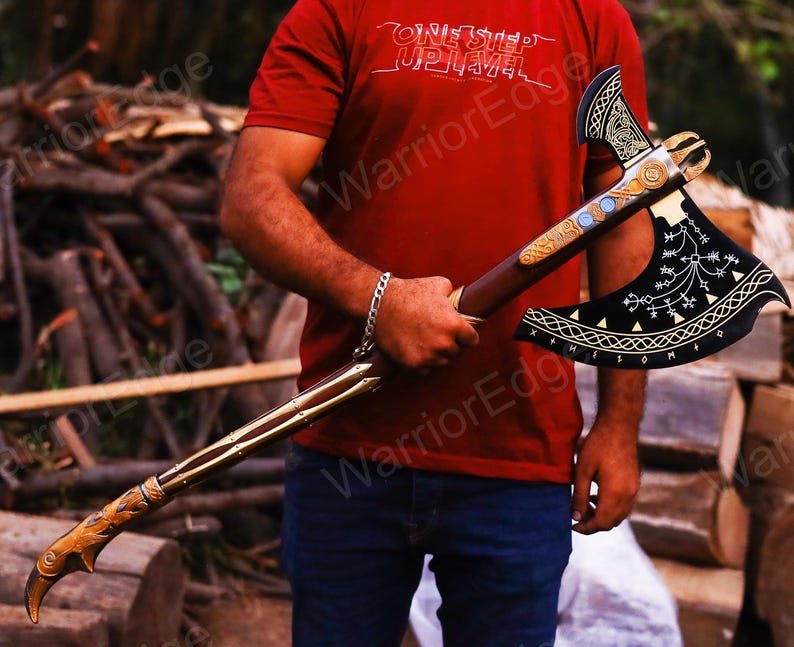 May include: A black axe with gold and silver accents. The axe head has a decorative design with runes and symbols. The axe handle is made of wood and has a leather wrap. The axe is held by a person wearing a red shirt with the text "One Step Up Level" and "Warrior Edge".