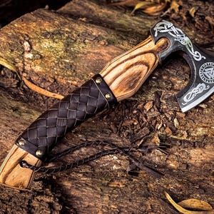May include: A black axe with a wooden handle and a leather wrap. The axe head is decorated with intricate carvings and a symbol of a triangle with a circle around it. The axe is resting on a piece of wood.