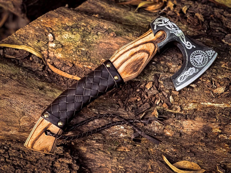 May include: A black axe with a wooden handle and a leather wrap. The axe head is decorated with intricate carvings and a symbol of a triangle with a circle around it. The axe is resting on a piece of wood.
