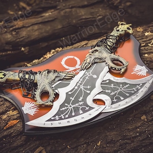 May include: A pair of black and silver double-bladed axes with intricate designs and a chain connecting them. The axes are displayed on a red and black background with the word "God of War" printed on it.