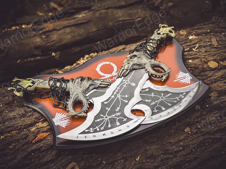 May include: A pair of black and silver double-bladed axes with intricate designs and a chain connecting them. The axes are displayed on a red and black background with the word "God of War" printed on it.