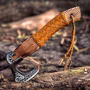 May include: A hand-forged axe with a leather-wrapped wooden handle and a braided leather strap. The axe head features intricate carvings and a decorative design.