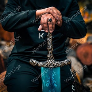 Pode incluir: Imagem detalhada de uma espada com uma l&acirc;mina azul e uma empunhadura ornamentada e texturizada. A empunhadura tem um design semelhante a uma corda com detalhes dourados. A espada &eacute; segurada por uma pessoa vestida de preto.