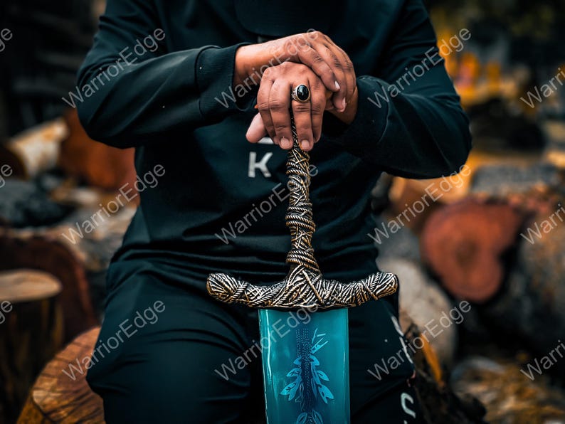 Pode incluir: Imagem detalhada de uma espada com uma l&acirc;mina azul e uma empunhadura ornamentada e texturizada. A empunhadura tem um design semelhante a uma corda com detalhes dourados. A espada &eacute; segurada por uma pessoa vestida de preto.