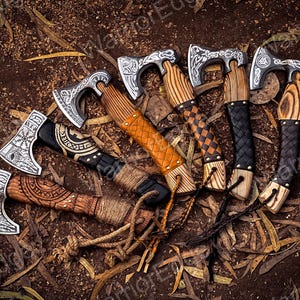 May include: A set of six hand-forged Viking axes with intricate carvings on the blades and wooden handles. The axes have leather wrappings and are arranged in a circle on a bed of brown leaves.