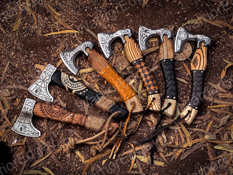 May include: A set of six hand-forged Viking axes with intricate carvings on the blades and wooden handles. The axes have leather wrappings and are arranged in a circle on a bed of brown leaves.