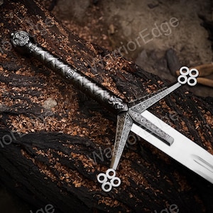 May include: A silver-toned metal sword featuring an intricate crossguard with four-ringed terminals and a textured, patterned grip. The pommel is detailed with embossed designs, and the blade is smooth, polished steel with a central fuller groove.