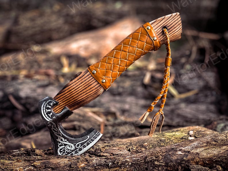 May include: A hand-forged axe with a leather-wrapped wooden handle and a braided leather strap. The axe head features intricate carvings and a decorative design.