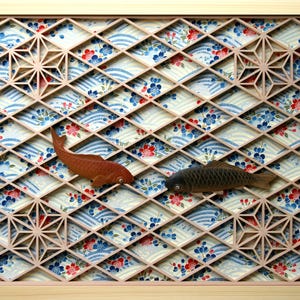 May include: A wooden frame with a geometric pattern and a floral background. Two wooden fish are placed within the frame, one brown and one black.