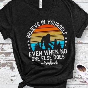 May include: Black t-shirt with a retro sunset graphic and the text "Believe in yourself even when no one else does - Bigfoot".