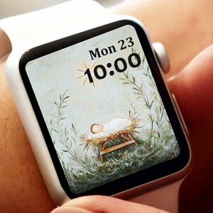 Peut inclure: Une montre connectée blanche affichant une image religieuse d'un bébé dans une crèche, entouré de verdure. L'écran affiche également la date, lundi 23, et l'heure, 10h00. La montre est portée au poignet, un doigt touchant l'écran.