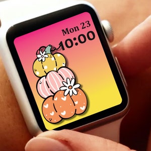 May include: A white smartwatch with a colorful pumpkin-themed watch face. The watch face displays three stacked pumpkins in yellow, pink, and orange, with white daisy accents. The time is 10:00 on Monday, the 23rd.