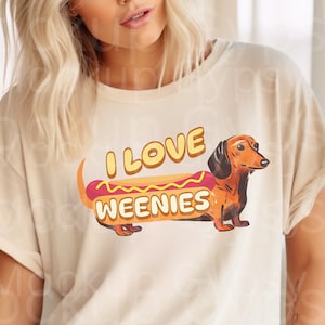 May include: A beige t-shirt featuring a dachshund dog dressed as a hot dog. The text on the shirt reads "I love weenies".