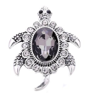 May include: A silver-toned turtle-shaped brooch with a large, oval, gray gemstone center. The brooch is embellished with clear, round crystals and detailed with textured patterns on the shell and flippers. The turtle's head features two small, black gemstones for eyes.