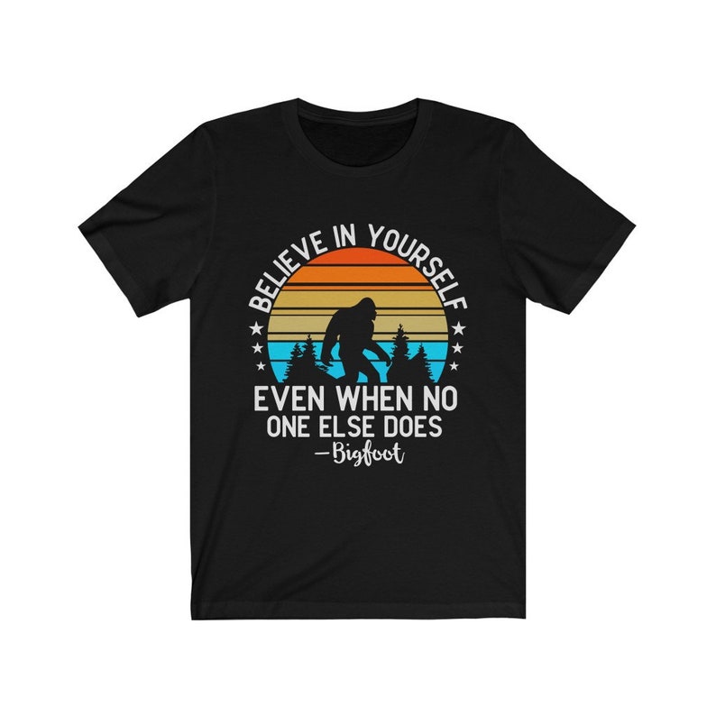 May include: Black t-shirt with a retro sunset graphic featuring a silhouette of Bigfoot. The text reads "Believe in yourself even when no one else does - Bigfoot".
