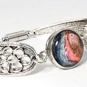 May include: A silver spoon bracelet with a floral design and a round, colorful, abstract resin charm. The bracelet has a magnetic clasp.