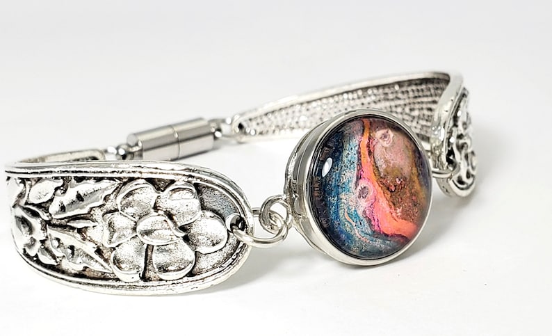 May include: A silver spoon bracelet with a floral design and a round, colorful, abstract resin charm. The bracelet has a magnetic clasp.
