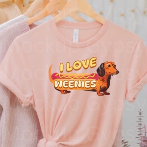 May include: A pink t-shirt featuring a dachshund dressed as a hot dog. The text on the shirt reads "I love weenies".