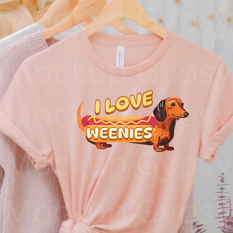 May include: A pink t-shirt featuring a dachshund dressed as a hot dog. The text on the shirt reads "I love weenies".