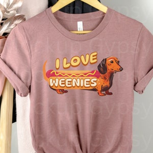 May include: A pink t-shirt featuring a dachshund dressed as a hot dog. The text on the shirt reads "I love weenies".