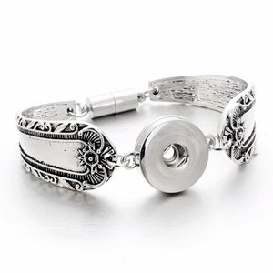 May include: Silver-toned bracelet featuring a unique design. The bracelet incorporates decorative spoon-shaped elements with ornate detailing. A large, round snap closure is centered, and a magnetic clasp secures the bracelet. The overall style is vintage-inspired.