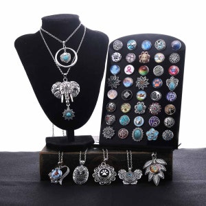May include: A set of silver charm necklaces with various pendants, including an elephant, owl, heart, paw print, and leaf. The necklaces are displayed on a black stand with a variety of colorful snap buttons.